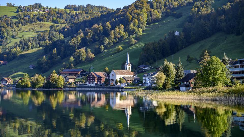 Die neuromanische Kirche spiegelt sich im Ägerisee. Umgeben wird der Sakralbau von Bäumen und Wiesen.