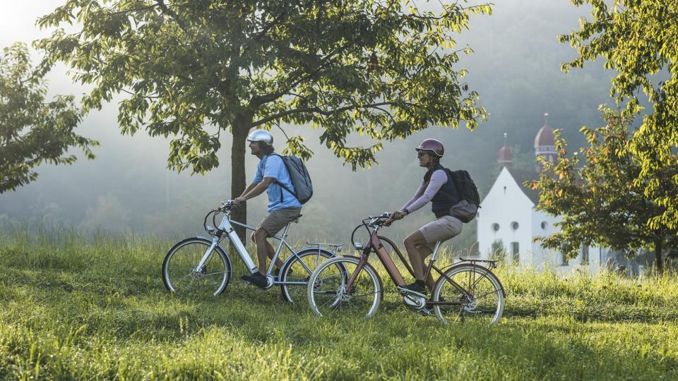 Eine Frau und eine Mann fahren auf dem Velo vor einer Kapelle durch. Die Kapelle steht auf einer Wiese und ist mit Kirschbäumen umgeben.