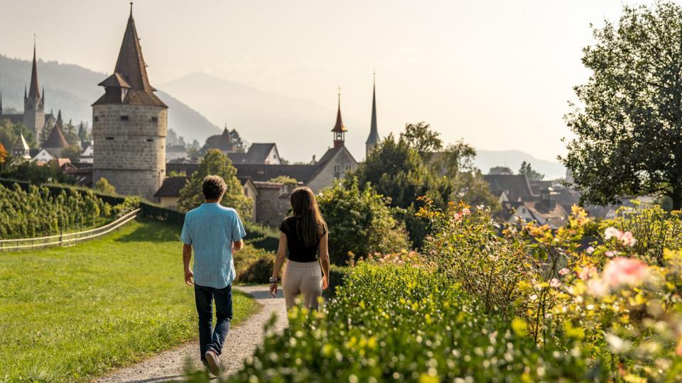 Zwei Menschen spazieren vor blühenden Rosen auf einem Kiesweg zu einem historischen Gebäude.