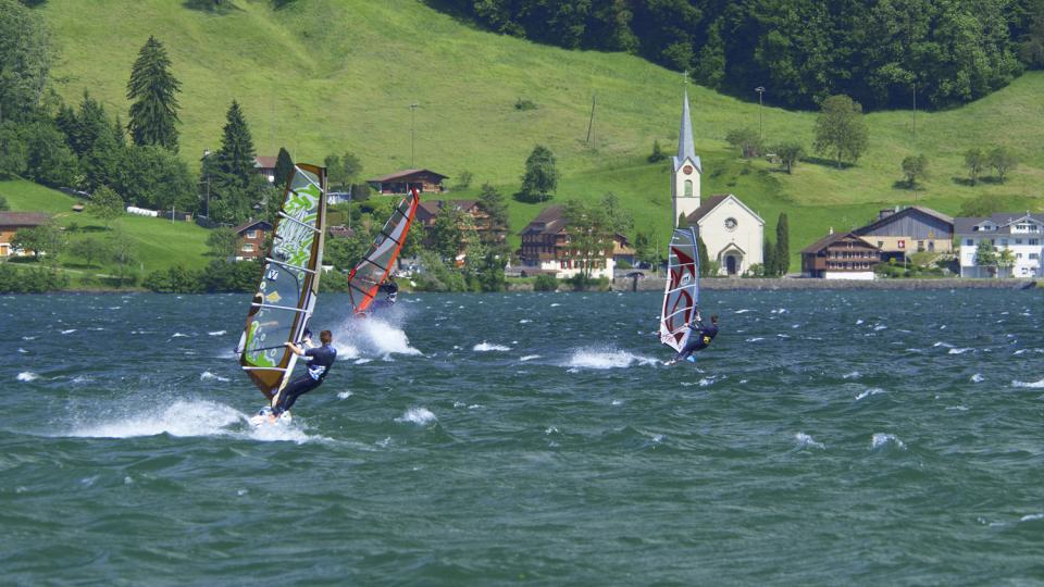 Drei Windsurfer surfen auf dem unruhigen Ägerisee. Im Hintergrund sind die Kirche und Holzhäuser zu sehen.