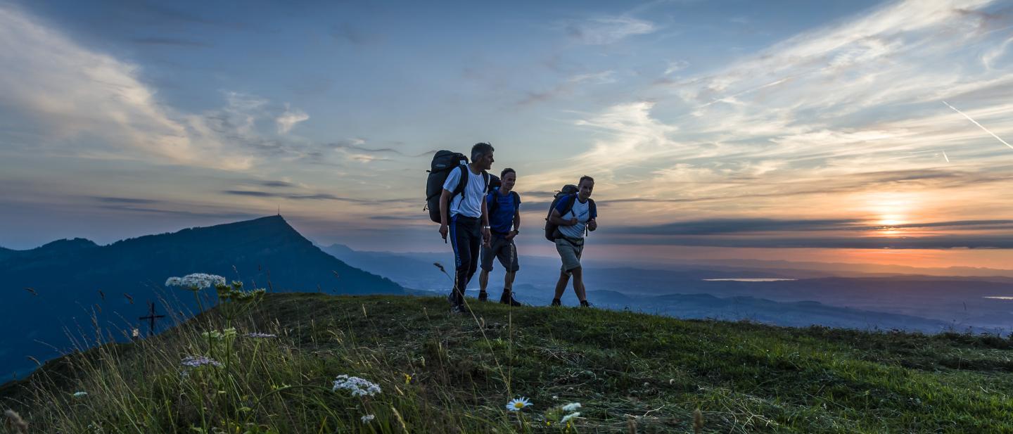 Drei Männer mit Gleitschirmrucksäcken wandern über den Gnipen. Im Hintergrund geht die Sonne unter,