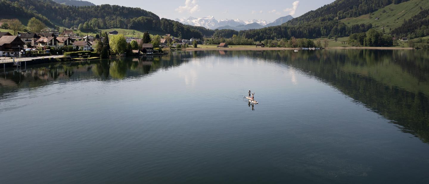 Zwei Stand-up-Paddelnde sind auf dem Ägerisee. Im Hintergrund sind die schneedeckten Gipfel der Alpen zu sehen.